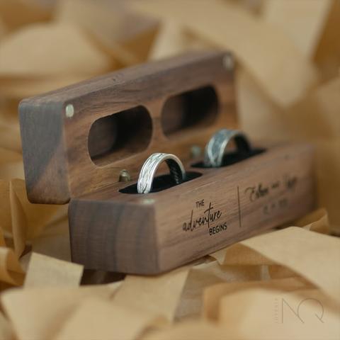 en un fondo de  papeles marrones una caja de madera con hueco para 2 alianzas.
las alianzas tienen tallados de formas tranzadas entremezcladas y mezclan plata blanca y envejecida