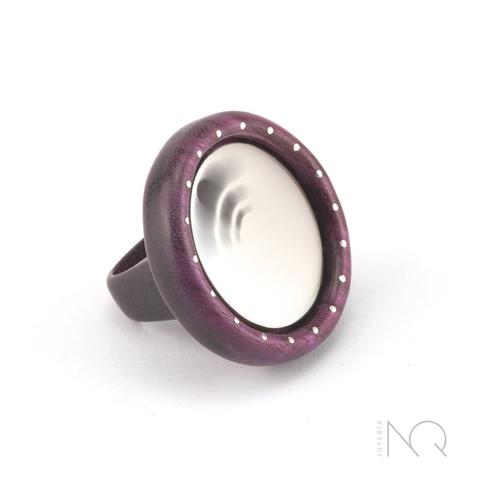 Foto de un anillo de madera pulida de tono morado, el brazo del anillo es grueso de madera y sostiene una forma circular también de madera, dentro de ella hay otro circulo de plata blanca con 3 agujeritos en una esquina.
en esta foto se ve como la parte central de plata se mueve girando y desdibujando los 3 agujeros circulares