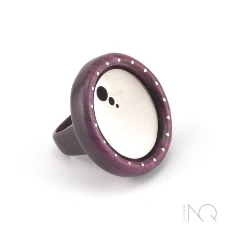 Foto de un anillo de madera pulida de tono morado, el brazo del anillo es grueso de madera y sostiene una forma circular también de madera, dentro de ella hay otro circulo de plata blanca con 3 agujeritos en una esquina.