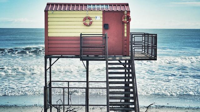 A red and yellow wooden lifeguard hut stands on stilts on the sandy Brittas Bay beach in County Wicklow, Ireland, with the blue Irish Sea and white breaking waves in the background under a bright sky. Two red lifebuoys are attached to the front of the hut. A metal staircase leads up to the entrance, and there are metal grated platforms extending from the hut.