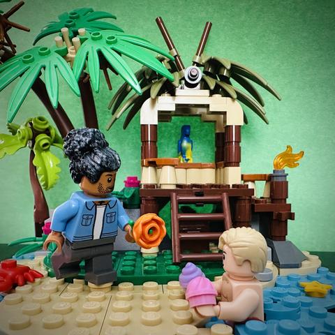 Lego Ed offering a flower to Lego MerStede, who is submerged in the water offering sea shells. They are at a beach, with palm trees and a small hut in the background.