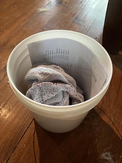Two gallon white plastic tub. In the tub are pattern pages and a knitting work in progress. The yarn is grey. Not visible under the wip is the large cone of yarn. It is all sitting on a wooden floor.