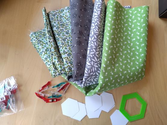 Four pieces of patterned fabric on green and grey, some fabric clips, hexagonal paper cutouts, and a green hexagon stencil