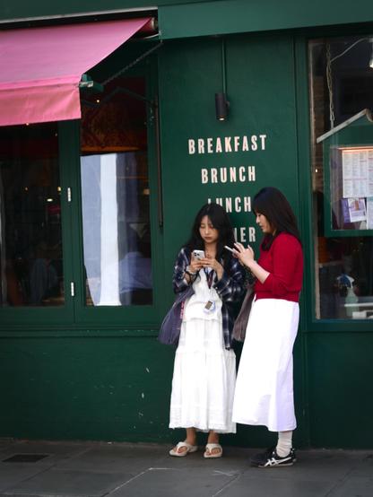 Two young dark-haired women who look like a mother and her teenage daughter, similarly dressed in white skirts, standing outside a green-painted central London cafe between two windows and in front of the words in this photo's title, both checking their phones.