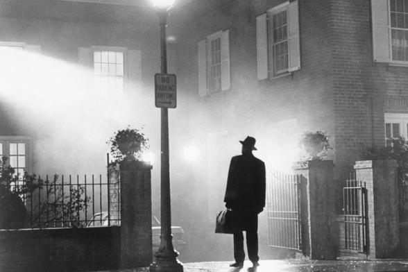 This is a still of an exorcist dressed in all black, with a fedora hat on, looking up at a house. He's standing under a straight lamp in the driveway.
