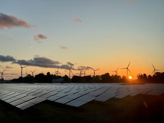 Sunset landscape, showing an orange sun about to set. Wind turbines silhouetted in the background, and in the foreground an array of solar panels.