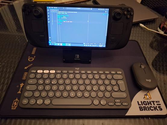 Auf einem Tisch steht ein Steam Deck auf einem Ständer, davor eine kleine Tastatur und eine Maus. Auf dem Display des Steam Deck läuft Visual Studio Code, es ist eine COBOL-Datei geöffnet.