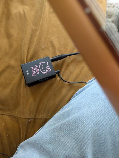 A small black Hello Kitty branded guitar headphone amp on a couch. It has a guitar cord plugged into it as well as headphones. There is an out of focus guitar neck in the foreground.