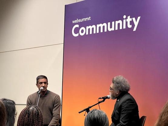 Ramesh Srinivasan and Cornell West sit at mics in conversation in front of a sign saying “WebSummit Community”