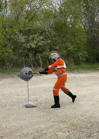 A person dressed as a Star Wars x-wing pilot in orange jumpsuit and helmet smacking a paper mache Death Star with a baseball bat.