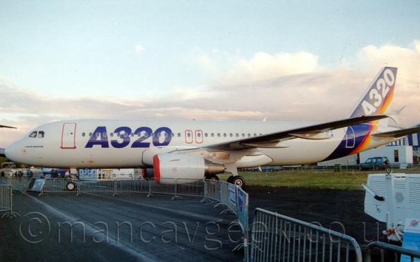 Side view of a tin-engined jet airliner parked facing to the left.
The plane is mostly white, with large, blue, billboard-style "A320" titles on the forward fuselage, and a thick blue bland, with thinner red, orange, and yellow bands, wrapped around the rear fuselage and tail, with white "A320" titles on the blue section.
The engine pods under the wings have red covers on the inlet.
The plane is parked on a black tarmac taxiway, behind a temporary metal fence which curves through the bottom of the frame to provide a walkway for spectators.
Fluffs of white cloud with hints of pale blue fill the rest of the frame.