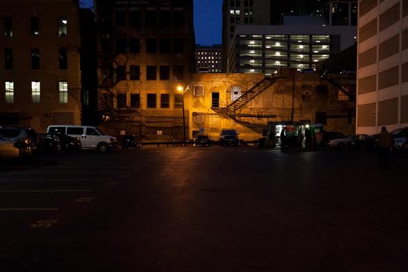 A nearly empty parking lot with buildings surrounding it at night. A single sodium lamp illuminates the back of the parking lot casting shadows and light on the nearby buildings. Further behind other buildings with lights in their windows are seen (save one, to the left of the sodium lamp)