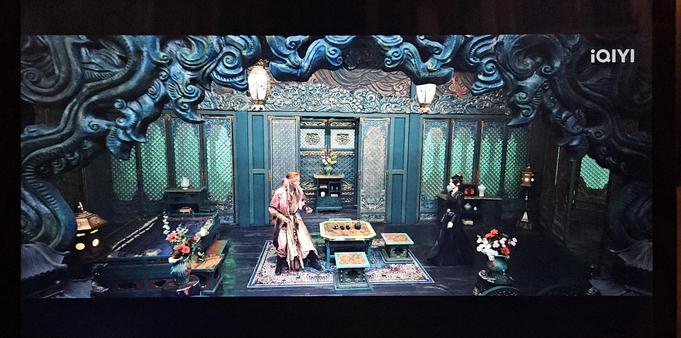 Mu Jiu jumping up from his stool in his room as Tian Huo walks straight towards him. The screen in the foreground is carved with decorative clouds, and the wall behind the actors  opens in the middle to display a storage unit with a flower vase. Moreover the wall have lattice work silk screen windows that lets in green light from the corridor.