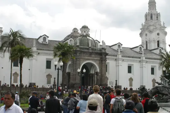 The image depicts a large historic building with a prominent architectural style, possibly located in a plaza or public space. There are many people gathered around the area, with some palm trees in the foreground. The building features a dome and detailed facades, suggesting it might be a significant cultural or governmental structure. The atmosphere appears lively, likely indicating a popular tourist destination.

Image Credits: Wikimedia / Quito magnífico / CC BY-SA 3.0