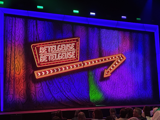 A theatre stage curtain with a neon sign that says “Betelgeuse Betelgeuse” on a backdrop of vibrant colours.