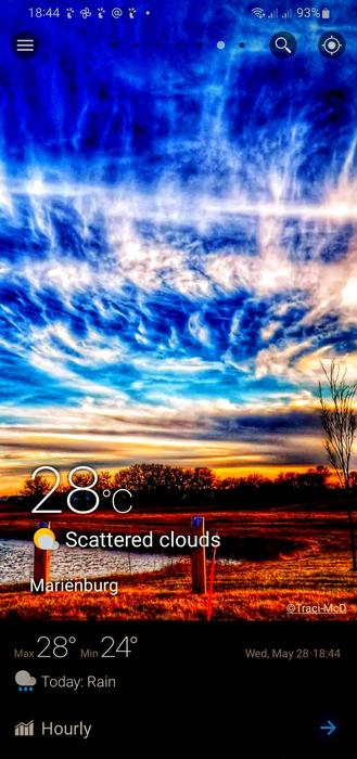 The image displays a smartphone screen with a weather app open. The background features a scenic image of a sunset with a dramatic sky, showcasing a mix of blue, white, and orange hues. The foreground includes a landscape with a body of water, grass, and a few trees, with a wooden post visible in the foreground.

The weather information is prominently displayed in the center of the screen. The current temperature is 28°C, with the condition noted as "Scattered clouds." The location is indicated as "Marienburg." The maximum temperature for the day is 28°C, and the minimum is 24°C. The weather forecast for the day includes rain. The date and time are shown as "Wed, May 28 18:44." Additional options such as "Hourly" are available at the bottom of the screen. The battery level is at 93%, and the time is 18:44.

Provided by @altbot, generated privately and locally using Ovis2-8B

🌱 Energy used: 0.233 Wh