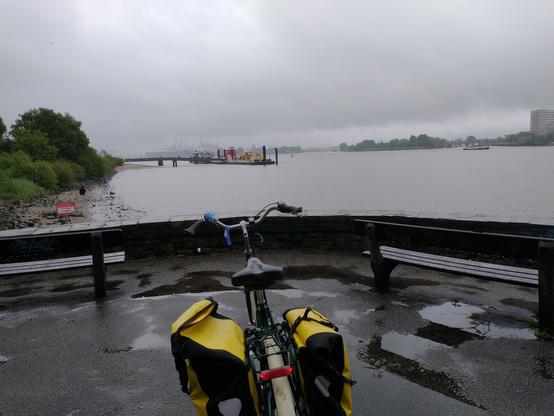 Ein Fahrrad mit gelben Taschen steht bei Bänken und einer kleinen Mauer. Dahinter die Elbe und der Fähranleger Teufelsbrück, an dem eine rote und eine gelbe Fähre liegen. Himmel und Wasser sind grau, das gegenüberliegende Ufer ist nur durch Dunst zu sehen.
