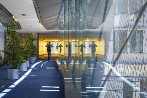 A man dressed in green is walking down a glass walkway towards an other pathway bathed in yellow light, in a public building in Brussels. His silhouette is multiplied by 5 on the glass walls.