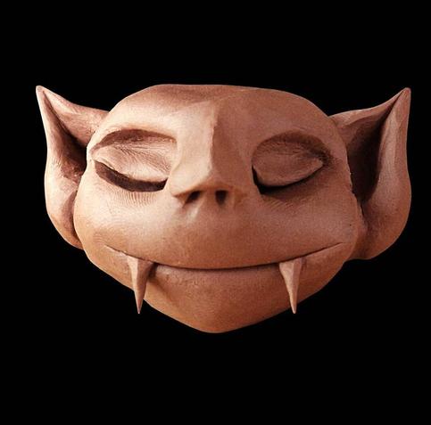 A plasticine sculpture of a stylized face with closed eyes and a gentle, content smile. The figure has pointed, elf-like ears and small protruding fangs or tusks from the lower jaw, giving it a whimsical, mythical creature appearance. The sculpture is rendered in warm terracotta or reddish-brown tones and photographed against a black background, creating dramatic contrast. The facial expression conveys serenity and peaceful satisfaction.