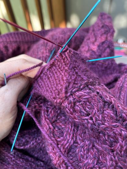 Tiny blue and red knitting nerds in a mulberry purple sweater cuff. A very pale human hand holds the thread and cuff in place