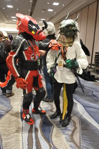 Fursuit photo - a dragon dressed in a nobles outfit poses next to a red Synth dragon (Xerian) at the dragon meet inside one of the convention spaces at Confuzzled 2025.  Photo was taken by Mutley