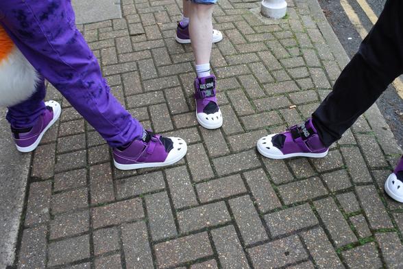 Photo of three furries at Confuzzled.  This is a shot of their shoes meeting in a triangle as they are happen to be wearing identical footwear.  In this case, the shoes are purple with paw markings on the front.
Left is a fox fursuiter, in the middle is his handler, and Hearts the lavender cat is to the right.