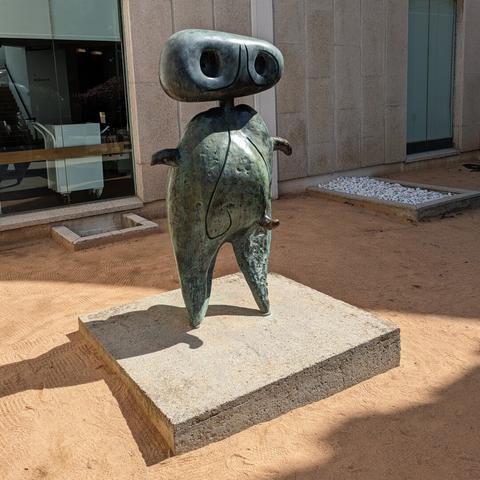 Joan Miró's "Personnage" sculpture is a tall, bronze figure that's both abstract and surreal. It's not a realistic depiction of a person but rather a whimsical, dreamlike form. Imagine a tall, vertical shape with a rounded head that's possibly incised with eyes and a nose. The surface has deep, gouged-out lines and three bulbous protrusions, suggesting a handmade quality. The figure evokes a sense of being both human and non-human, like a tree, root, or something entirely imaginary.