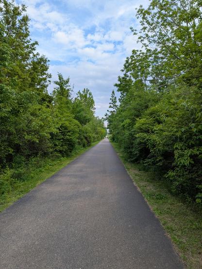 An asphalt paved path stretches out into the vanishing point lined with green trees and shubbery on both sides under a fairly cloudy sku with patches of blue peeking through.