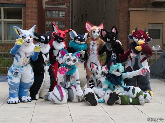 Fursuiters of various colors and species on a rooftop patio, in front of two brick facade buildings with an alleyway in between. There are six fursuiters standing in the back row, plus one kneeling, and two sitting on the ground in front.