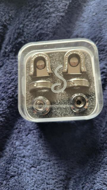 An image of Ruthenium S-Locks in the packaging. The box is square with rounded corners and made of clear plastic. There is a "Schaller" stylized S etched into the clear lid. 

There are two columns and three rows. Each corresponding row has identical parts. At the top is the horseshoe-shaped lock itself. In the middle are the screw thread locking wheels, which used to be standard hex nuts. At the bottom are the replacement strap buttons, with a hex screw embedded inside. 

The background is dark blue and fuzzy.
