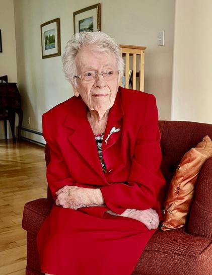 A white woman with white hair wearing a red jacket and skirt looking to the right of the photo, sitting on an orange chair with orange cushion, with a cream background. Photo taken on a Sunday morning in September in 2023 when she was 101.