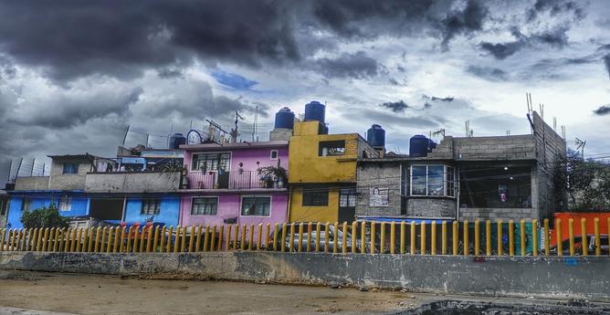 Tras una valla de postes amarillos de metal, una fila de casas, disparejas y multicolores, en un barrio pobre de la ciudad de México. Detrás de ellas, un cielo nublado empieza a oscurecerse.