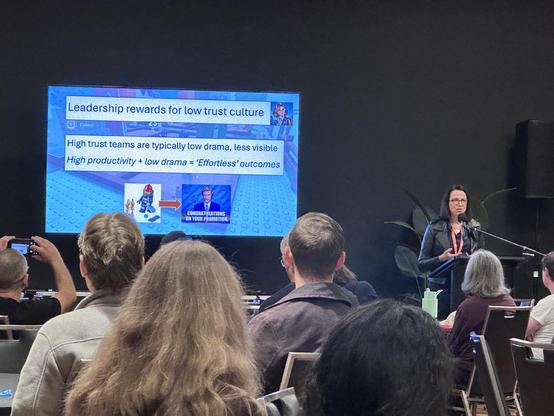 A woman presenting at a tech event with a slide that says:
Leadership rewards for low trust culture

High trust teams are typically low drama, less visible
High productivity + low drama = 'Effortless' outcomes