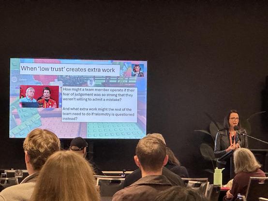 A woman presenting at a tech event with a slide that says:
When 'low trust' creates extra work

How might a team member operate if their fear of judgement was so strong that they weren't willing to admit a mistake?
And what extra work might the rest of the team need to do if telemetry is questioned instead?