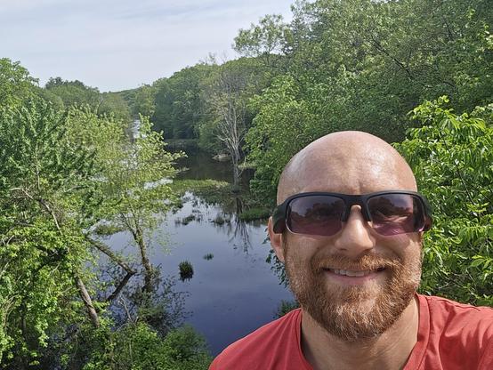 A selfie of me in front of a flooded swamp leading into the Charles River on a sunny day. Trees are growing out of the water, and thick forest lines both banks. I'm a bald, middle-aged, white man with a red beard flecked with white. I'm wearing black sunglasses and a red running shirt.