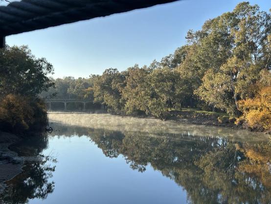View of the dark Murray River with a band of white mist being blown across it in the cold early morning. A bridge can just been seen midway & native trees are lining the bank against a beautiful blue sky.