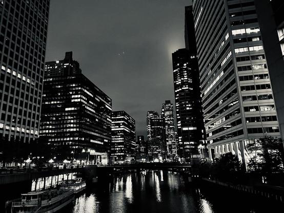 Black-and-white photo of buildings around the Chicago River, lights reflecting in the water.