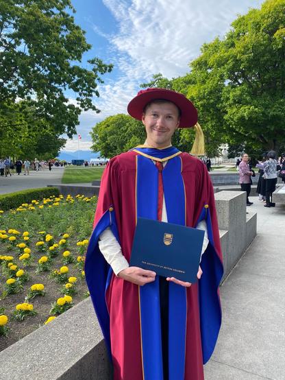 A man wearing a red and blue doctoral gown and a floppy red cap with a gold tassel stands in front of a flowerbed.