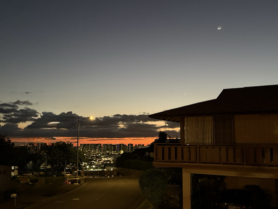 The last bit of sunset over Honolulu. Gray sky, clouds and orange under the cloud line. Crescent moon in the upper right of the picture.
