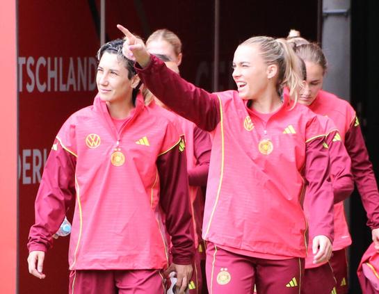 Sara Doorsoun und Laura Freigang kommen in pinken DFB-Trainingsanzügen aus dem Kabinentunnel, Freigang zeigt grinsend mit ausgestrecktem Arm nach links oben, Doorsoun guckt lächelnd in die Richtung, hinter ihnen sind weitere Spielerinnen. IMAGO/Jan Huebner.