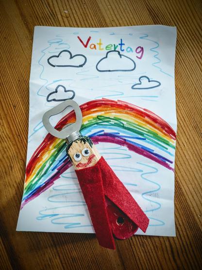 The image shows a handmade Father's Day gift. It features a drawing on a piece of white paper with the word "Vatertag" (German for Father's Day) written in colorful letters at the top. The drawing includes a large rainbow and several clouds. Attached to the paper is a bottle opener that has been decorated to look like a person with googly eyes, drawn-on hair and a mouth, and a red felt cape. The entire arrangement is placed on a wooden surface.