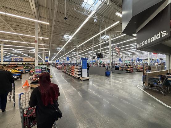 Der Blick geht vom Eingang eines Supermarkts in die riesige Halle mit vielen Regalen. Die Halle ist sehr hoch und mit langen Reihen von Neonlichtern beleuchtet. Im Vordergrund Kunden, rechts angeschnitten eine McDonald’s Filiale. Verglichen mit deutschen Supermärkten wirkt alles um ein vielfaches größer.