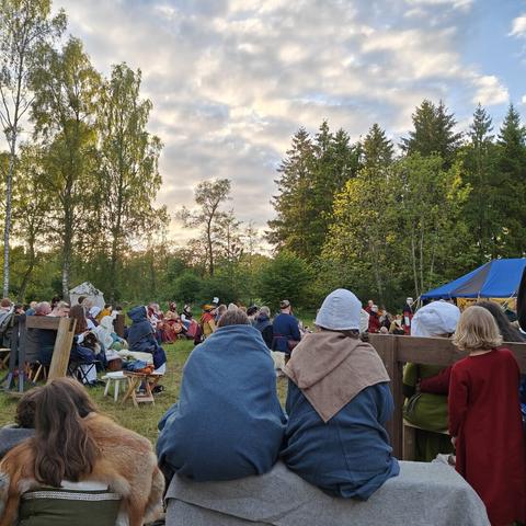 People in medieval clothing sitting on a field. In the background are green trees and a setting sun