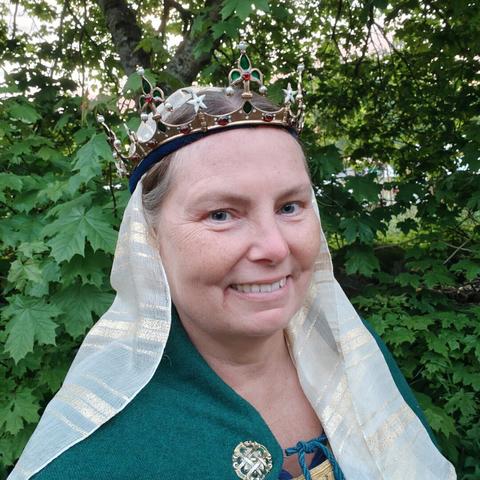 The head of smiling white middle aged woman wearing a green cloak, a white veil with gold stripes, an a crown, standing in front of a tree.