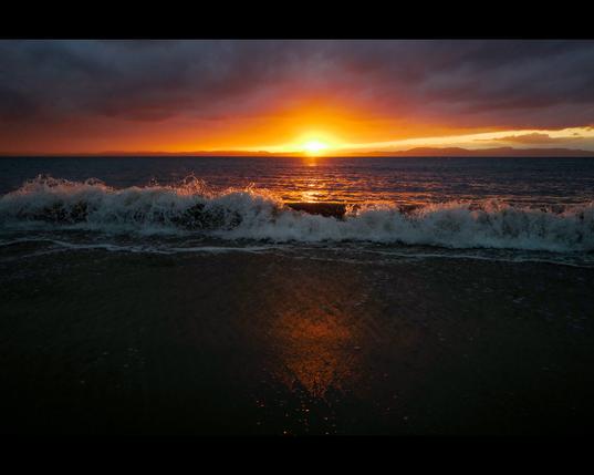 A sunset shot over the Dornoch Firth from Portmahomack with a breaking wave in the foreground. The warm sunlight reflected in the water and highlighting the spray from the top of the wave.