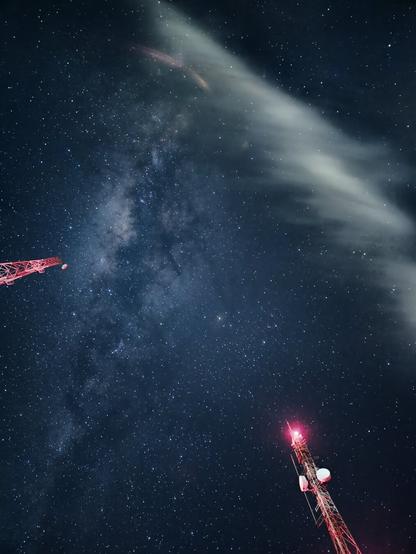 Upward-looking astrophotograph taken with a long exposure from Mirante Serra do Elefante in Mateus Leme, Minas Gerais, Brazil, using a Samsung Galaxy S23+. The image displays a brilliant night sky densely packed with stars, appearing as streaks or elongated points due to the long exposure. A discernible glowing band suggestive of the Milky Way stretches diagonally. The tops of two red-lit communication towers frame the bottom left and right corners, their structures angled inwards towards the impressive starscape. A soft streak of cloud crosses the upper portion of the frame, also showing signs of movement from the long exposure.