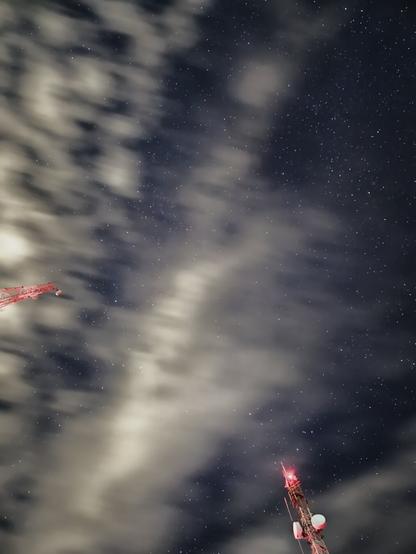 Astrophotograph captured with a long exposure from Mirante Serra do Elefante in Mateus Leme, Minas Gerais, Brazil, with a Samsung Galaxy S23+. The scene shows a vast expanse of stars exhibiting motion blur due to the long exposure, partially obscured by wispy, motion-blurred clouds also characteristic of the technique. Two red-lit communication towers are visible: a larger one with disc antennas in the lower right, and the tip of another appearing on the middle left edge of the frame.