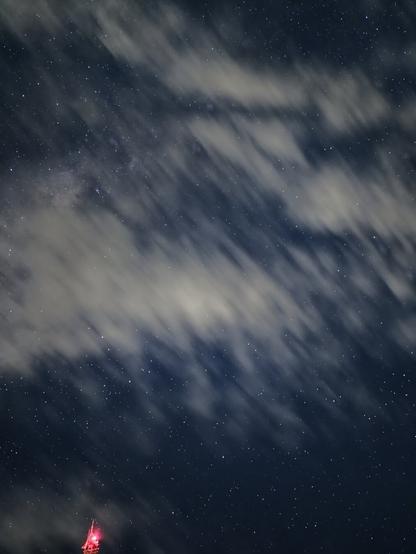 Night sky astrophotograph taken with a long exposure from Mirante Serra do Elefante in Mateus Leme, Minas Gerais, Brazil, using a Samsung Galaxy S23+. The image reveals numerous stars blurred by the Earth's rotation during the long exposure, and soft, elongated white clouds streaking diagonally across much of the frame. The bright red light from the top of a transmission tower is just visible in the extreme lower-left corner.