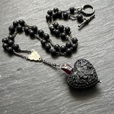 A photo of a rosary-style necklace on a dark grey slate background. The necklace chain is made from vintage black wooden rosary beads. The delicately carved cinnabar heart pendant is crowned with a red raw garnet nugget. The toggle clasp closure in made of pewter.