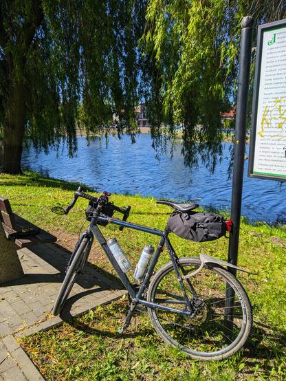 Ciemnoniebieski rower szutrowy oparty o słupek tablicy informacyjnej nad brzegiem miejskiego stawu. Pod siedzeniem z tyłu wystaje spora torba.